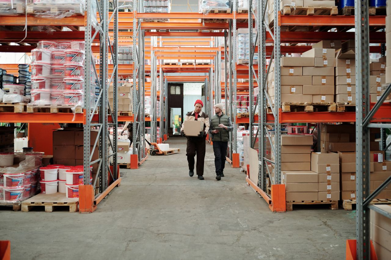 Services Two workers handle a package in a spacious warehouse surrounded by shelves stocked with boxes and products.